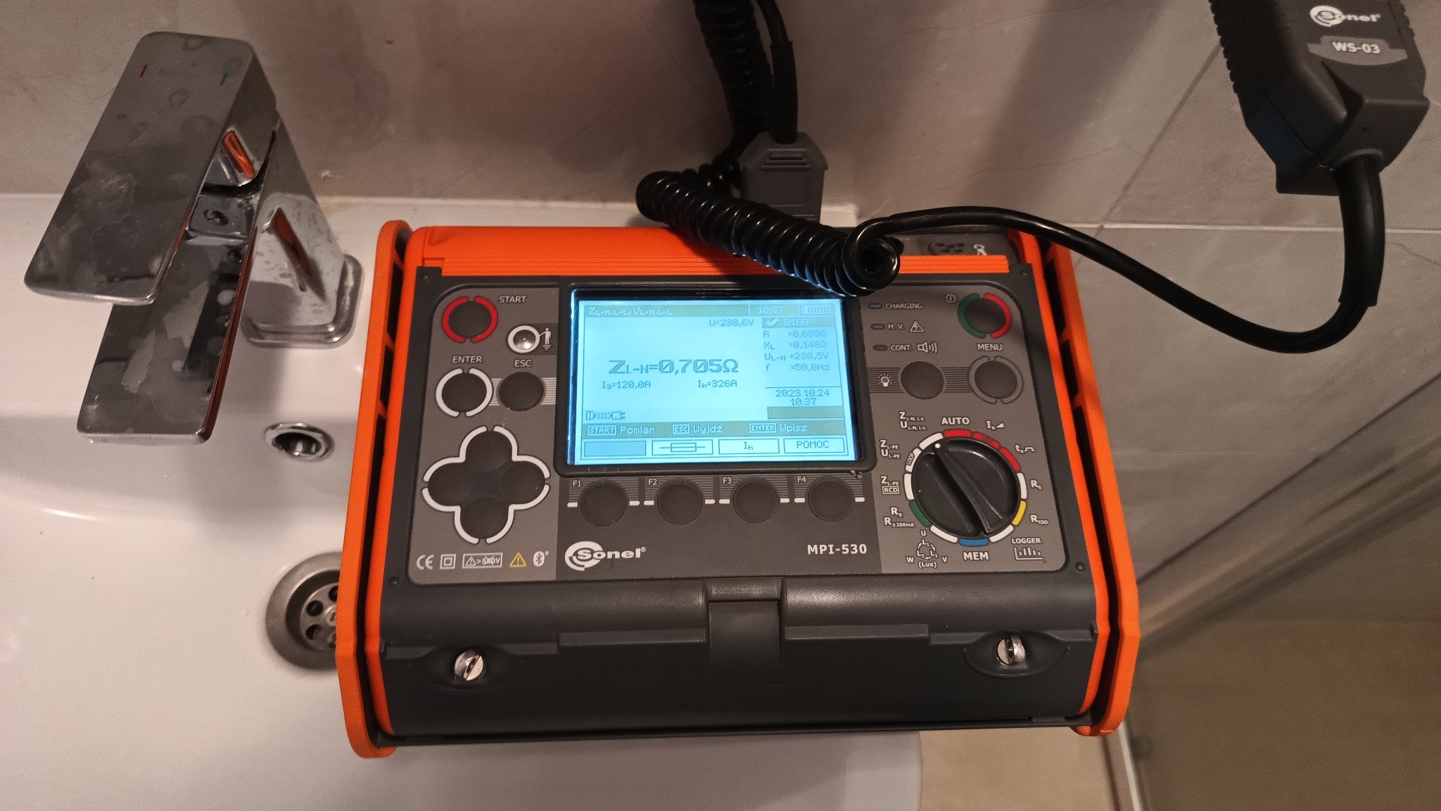 Miernik Sonel MPI-530 z wyświetlanymi wynikami pomiaru impedancji pętli zwarcia ZL-N=0,705Ω, leżący na umywalce w łazience.