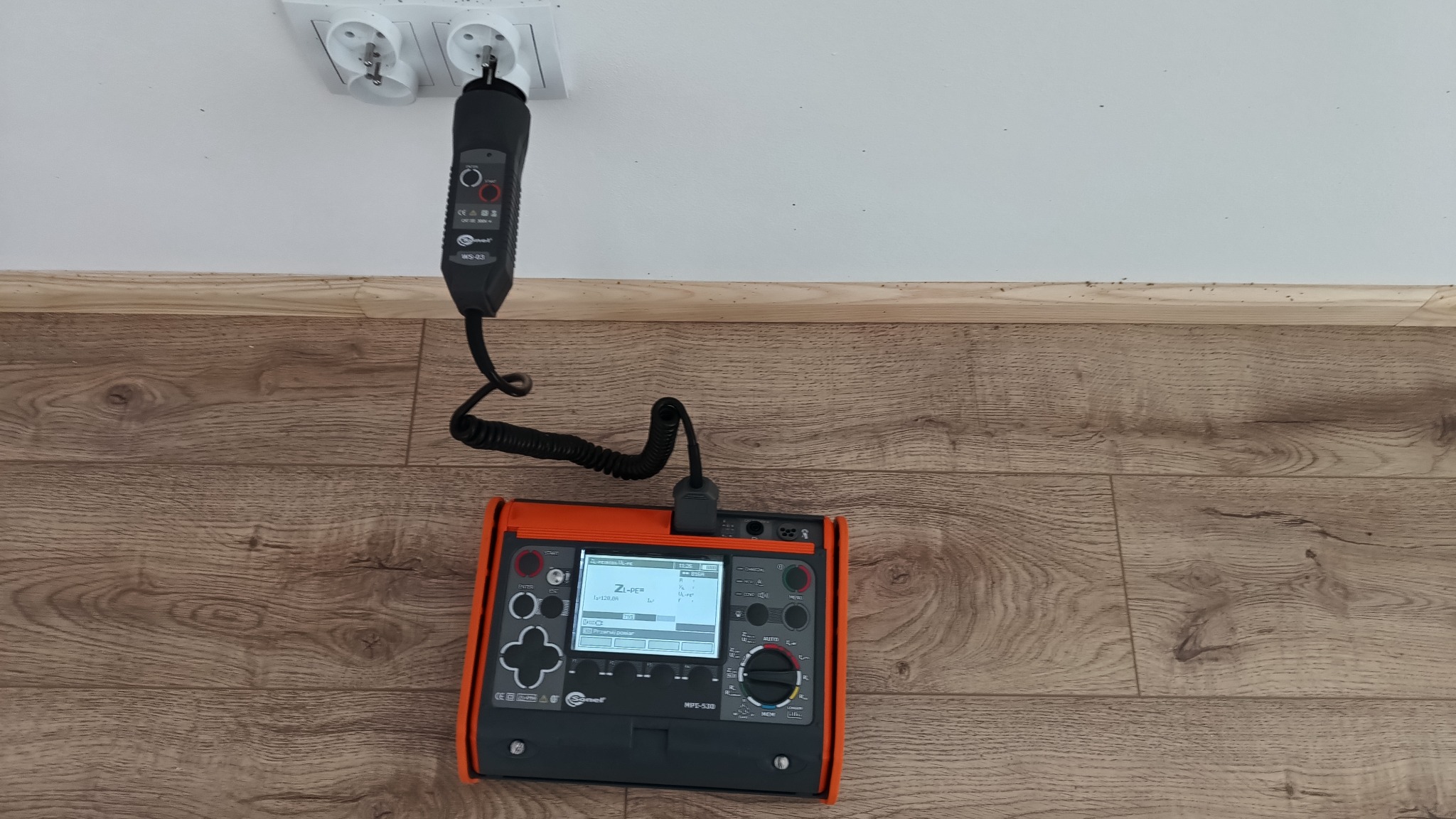 Miernik Sonel MPE-530 z podłączonym adapterem w gniazdku elektrycznym, leżący na podłodze z paneli.