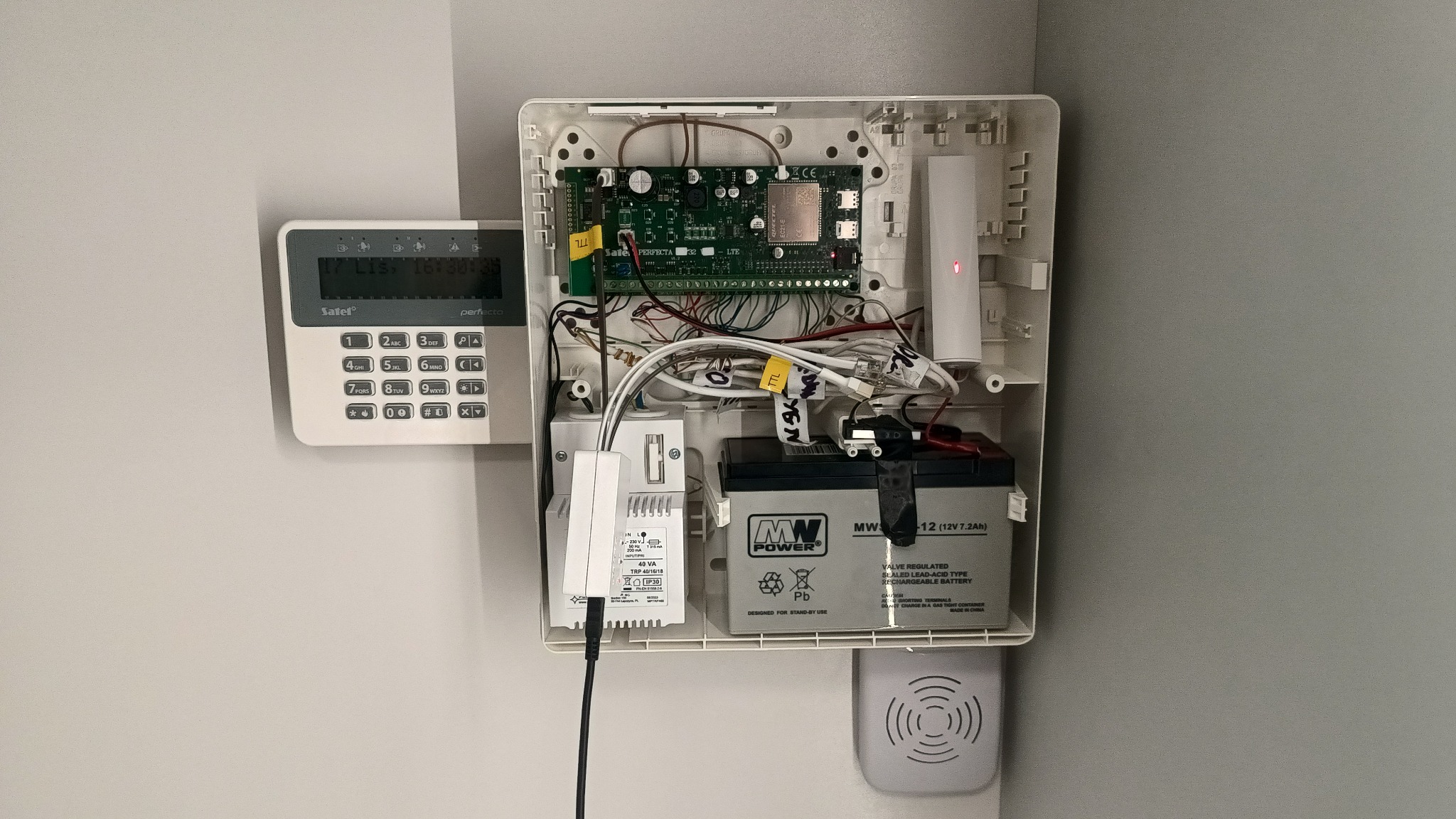 Otwarta obudowa centrali alarmowej Satel Perfecta LTE z widoczną elektroniką, akumulatorem MW Power 12V 7.2Ah, manipulatorem i sygnalizatorem akustycznym na szarej ścianie.