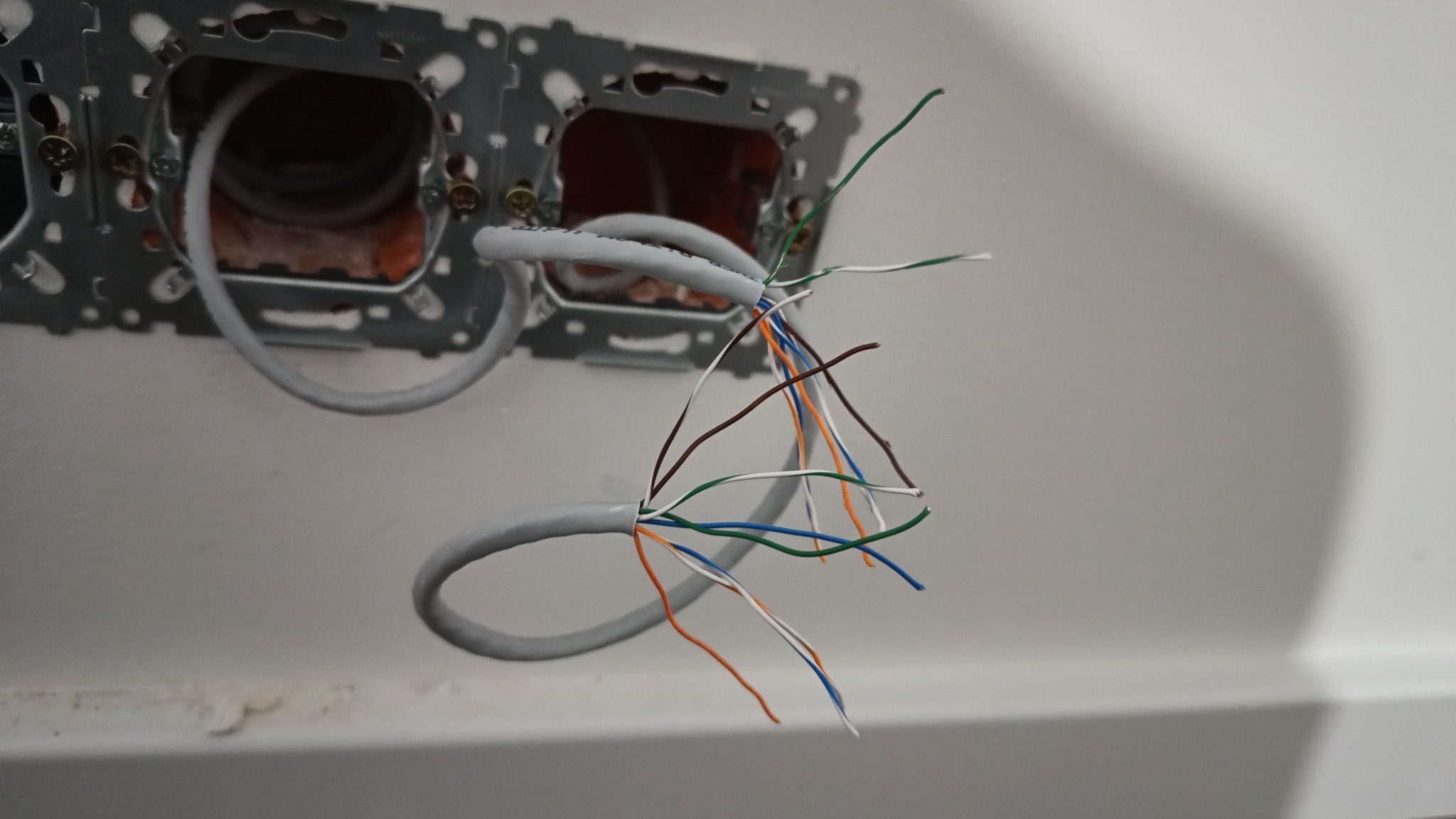 Szary kabel sieciowy z rozplątanymi kolorowymi żyłami wystającymi ze ściany, widoczne metalowe puszki montażowe gniazdek.