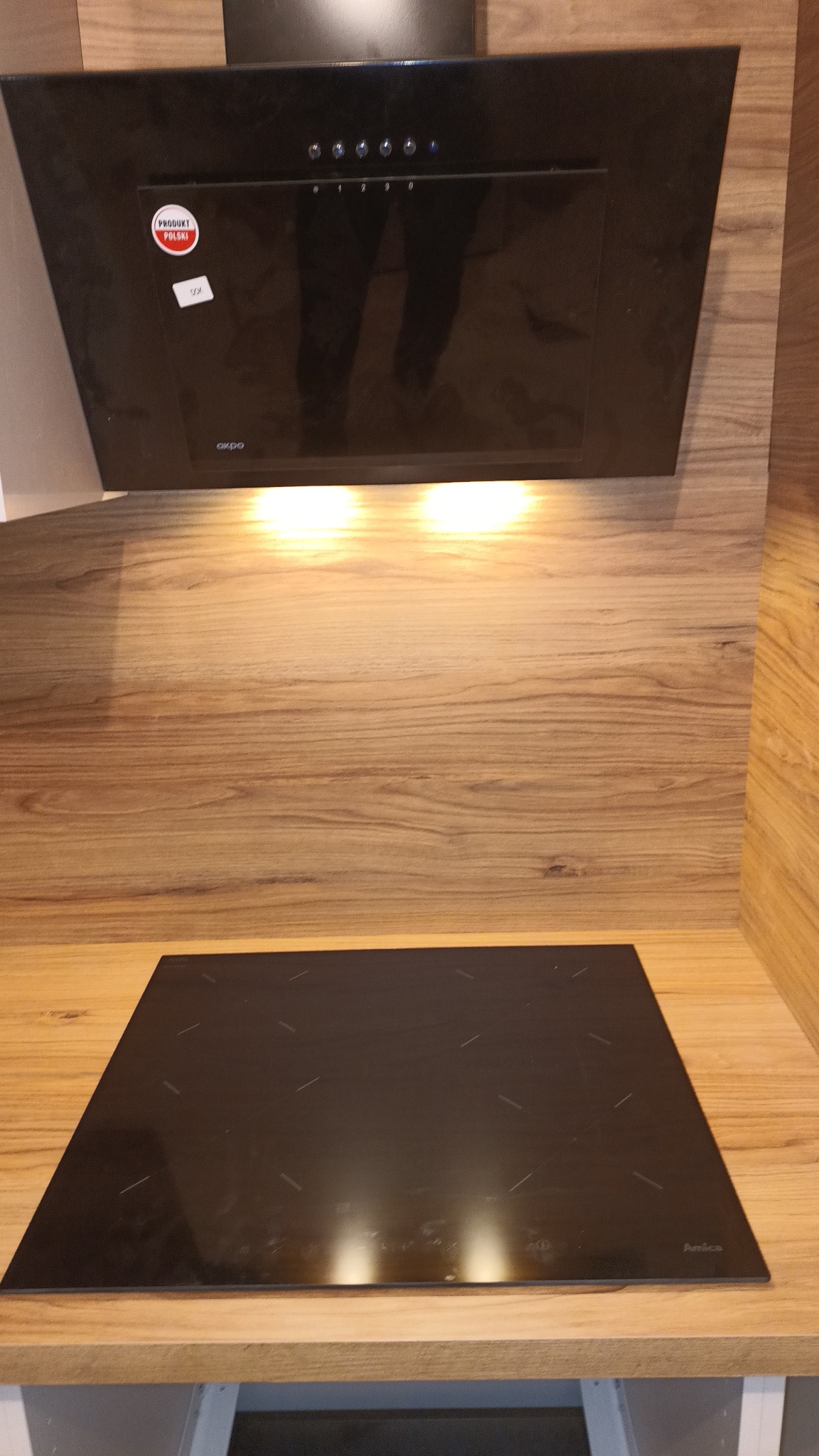 Czarna płyta indukcyjna marki Amica i okap kuchenny Akpo z naklejką 'Produkt Polski' zamontowane na drewnianym blacie.