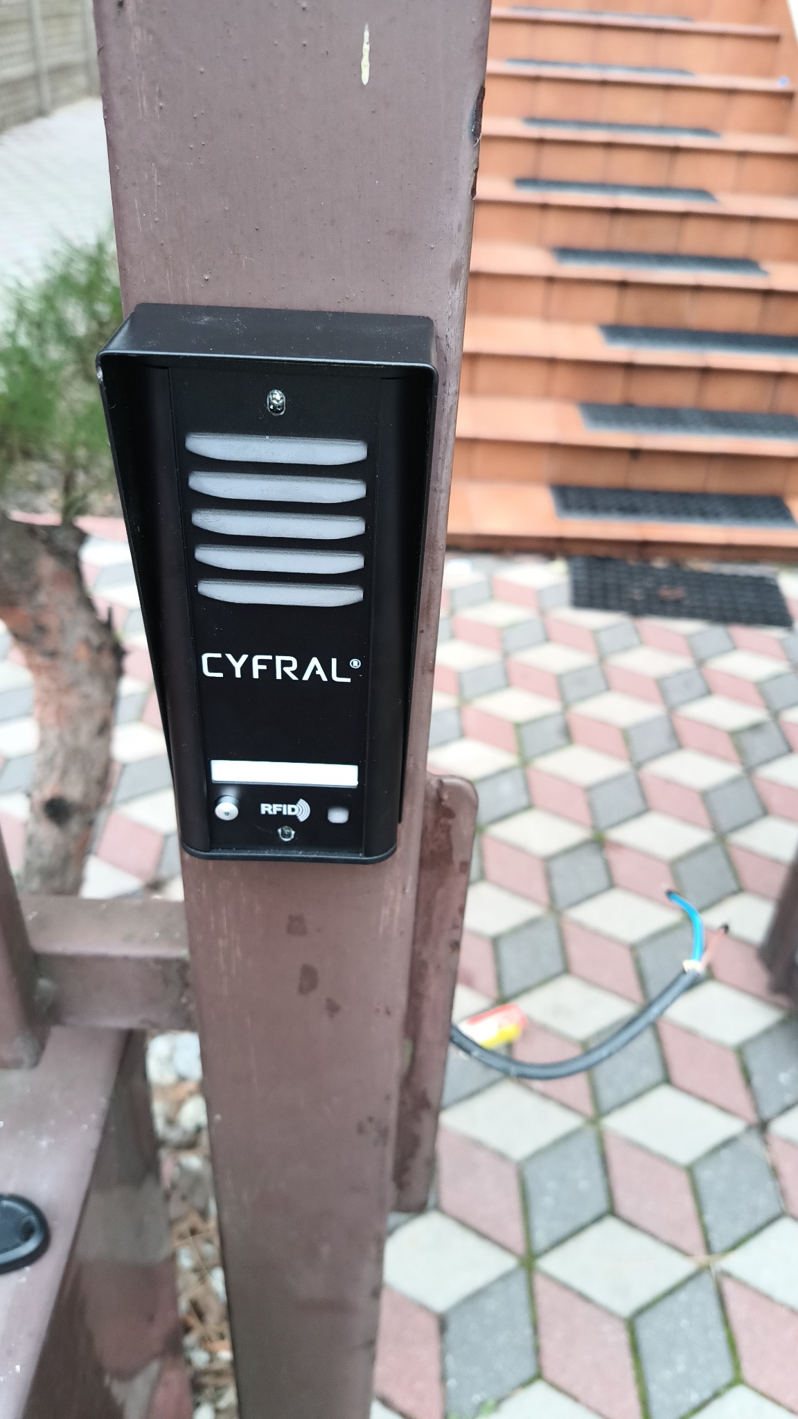 Czarny domofon Cyfral z czytnikiem RFID zamontowany na metalowym, brązowym słupku przy schodach wejściowych i kostce brukowej o geometrycznym wzorze. Widoczne wystające przewody.