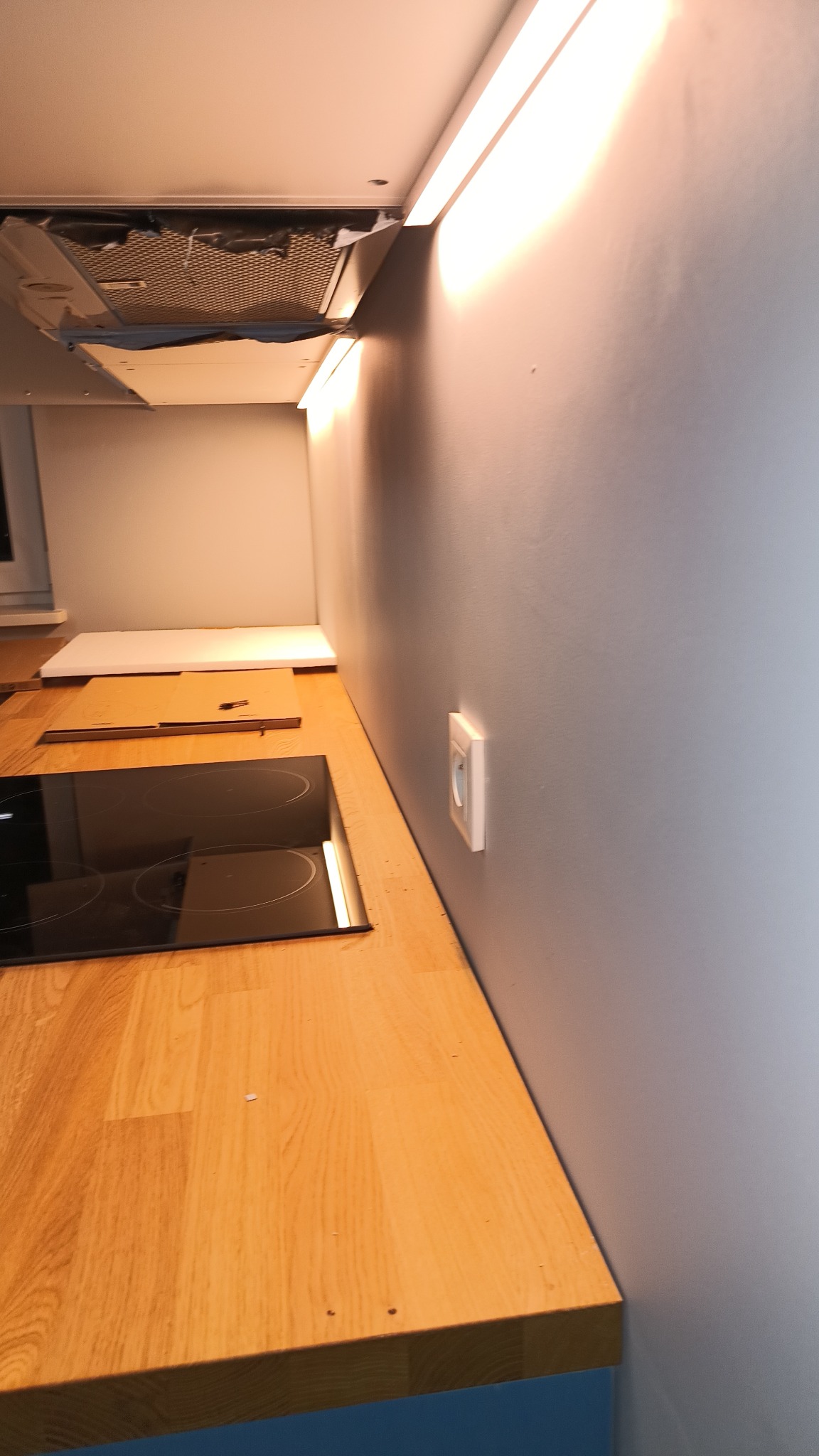 Częściowo zamontowana płyta indukcyjna na drewnianym blacie kuchennym, widoczny okap i gniazdko elektryczne na szarej ścianie, oświetlenie podszafkowe włączone.