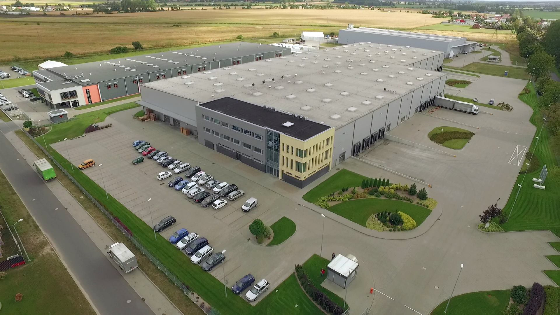 Szeroki kadr z lotu ptaka na kompleks magazynowy z rampami załadunkowymi, parkingiem pełnym samochodów osobowych i ciężarowych oraz zadbaną zielenią wokół, w tle rozległe pola uprawne.