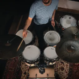 Perkusista w pasiastej koszulce gra na perkusji z talerzami, widok z góry na wzorzystym dywanie.