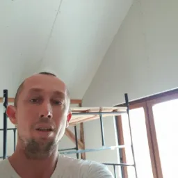 Mężczyzna w białej koszulce robiący selfie na tle sufitu z płyt gipsowo-kartonowych, widoczne ślady po wkrętach i rusztowanie.