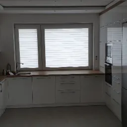 Nowoczesna kuchnia z białymi, błyszczącymi szafkami, drewnianym blatem i szarymi płytkami podłogowymi, oświetlona światłem dziennym wpadającym przez okno z żaluzjami.