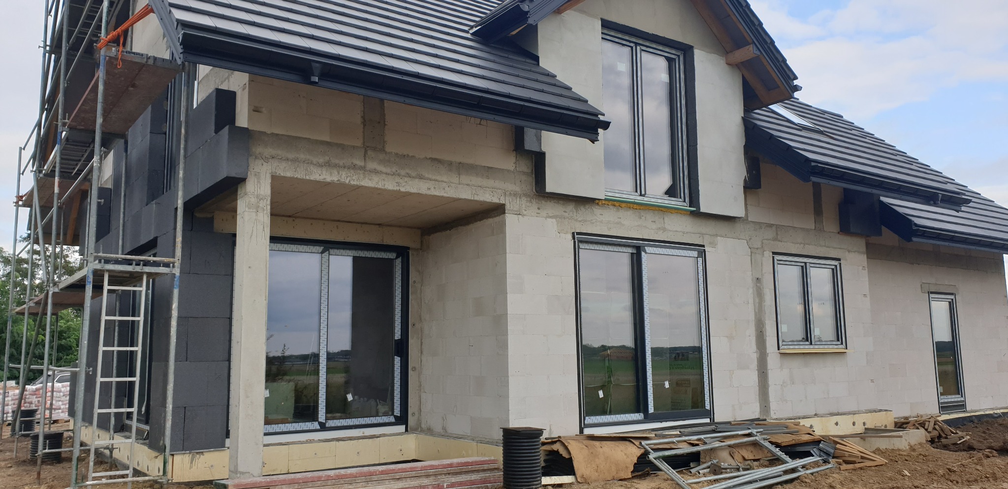 Dom w trakcie ocieplania: czarny styropian na elewacji, rusztowania, okna z czarnymi ramami, dach pokryty czarną dachówką.