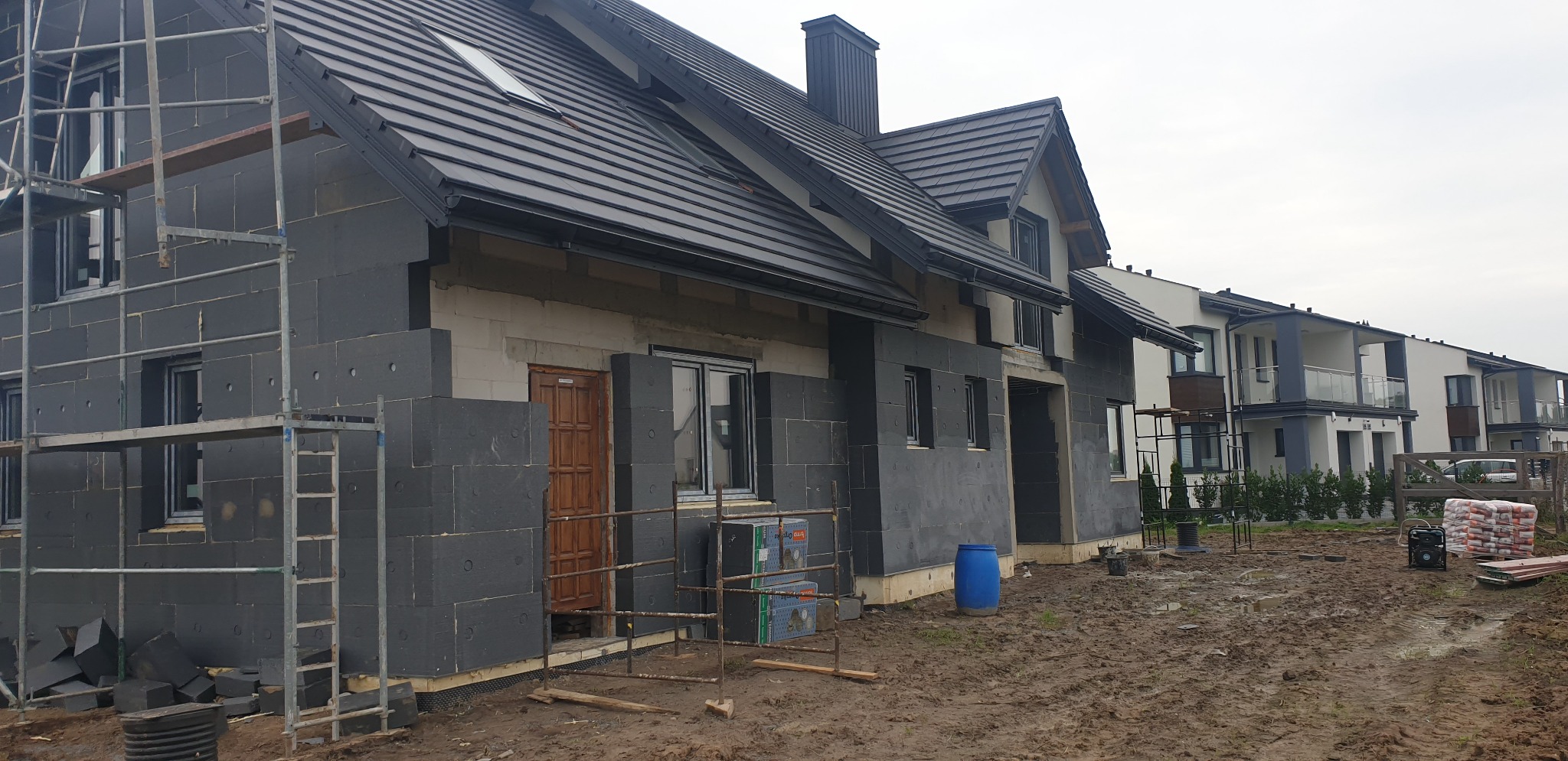 Dom w trakcie ocieplania, ściany pokryte czarnym styropianem, widoczne rusztowania i teren budowy w błocie.