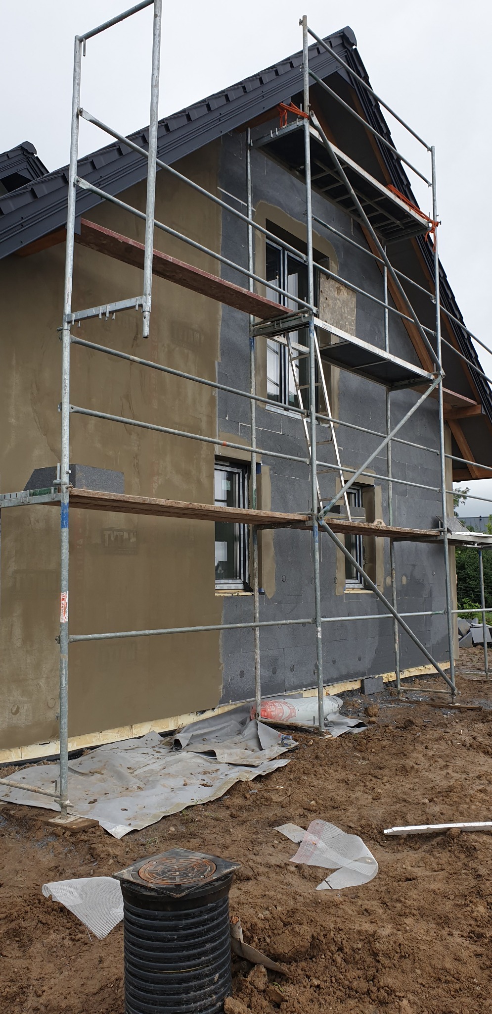 Dom w trakcie prac elewacyjnych: część ściany ocieplona styropianem, druga pokryta warstwą szarej zaprawy, rusztowanie metalowe, teren wokół rozkopany i zabezpieczony folią.