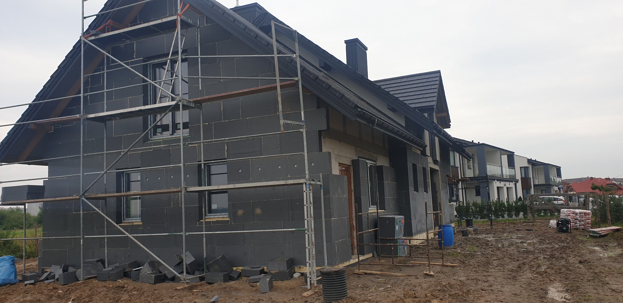 Dom w trakcie ocieplania, elewacja pokryta szarym styropianem, widoczne rusztowania i porozrzucane fragmenty izolacji, teren wokół rozkopany.