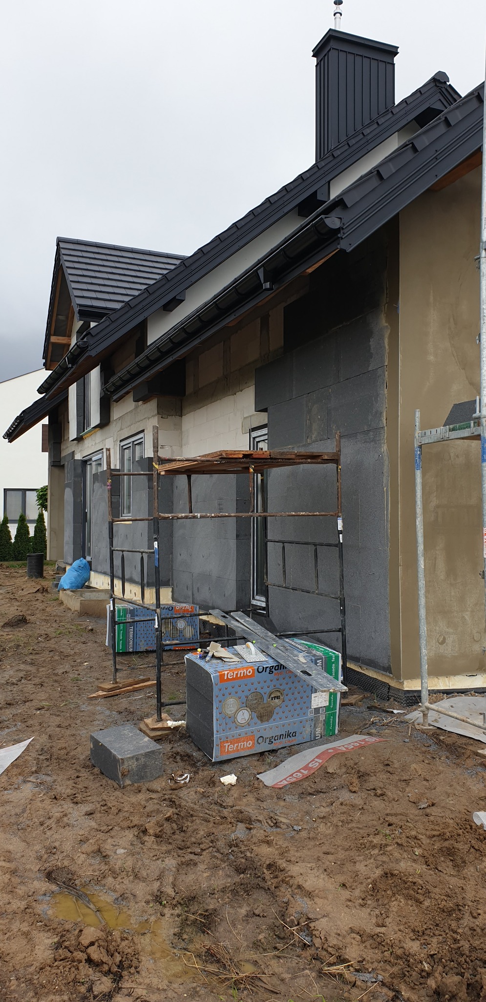 Dom w trakcie ocieplania: szare płyty termoizolacyjne na elewacji, częściowo otynkowana ściana, rusztowania, paczki styropianu Termo Organika na błotnistym podłożu.