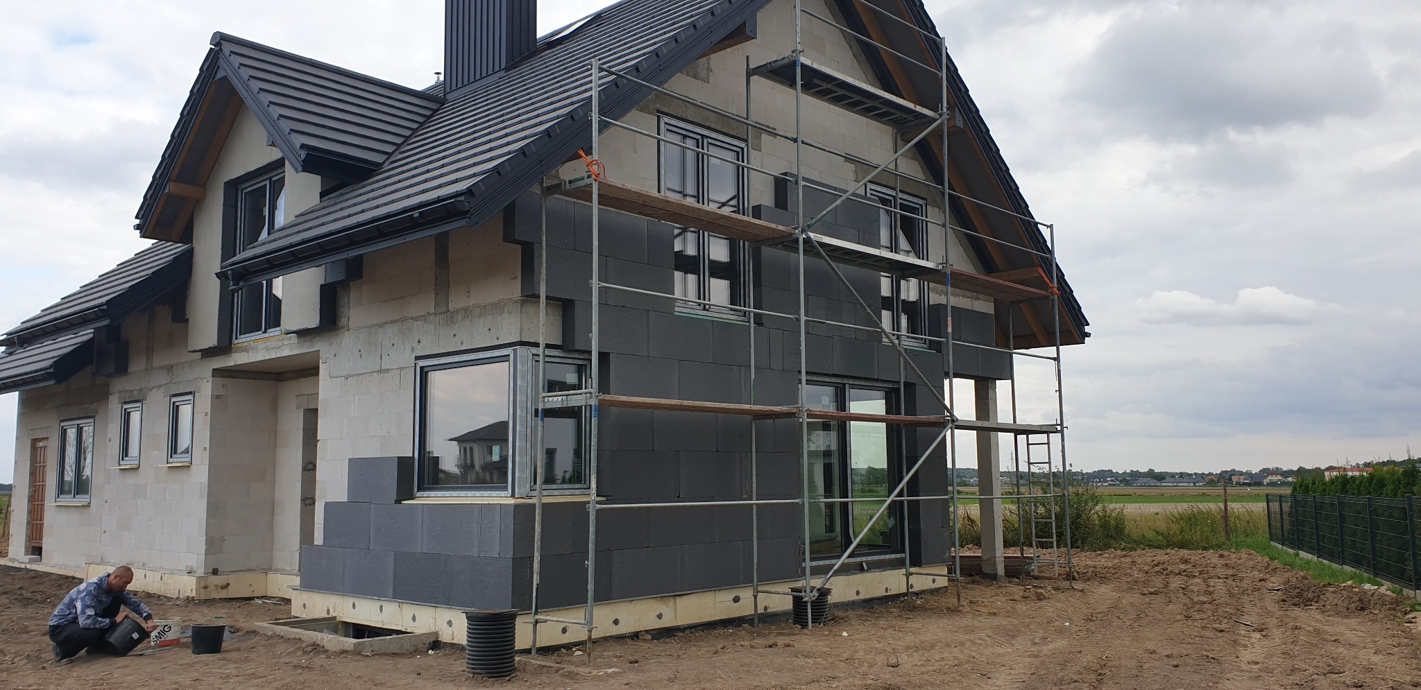 Dom w trakcie ocieplania, widoczne rusztowania, częściowo ocieplona ściana grafitowym styropianem, pracownik przygotowuje klej w wiadrze.