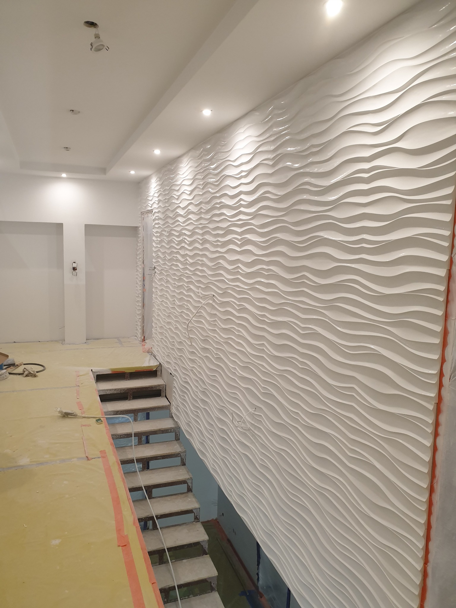 Biała ściana dekoracyjna z efektem fal, zamontowana w nowoczesnym wnętrzu obok schodów, z widocznymi zabezpieczeniami podłogi i surowym oświetleniem.