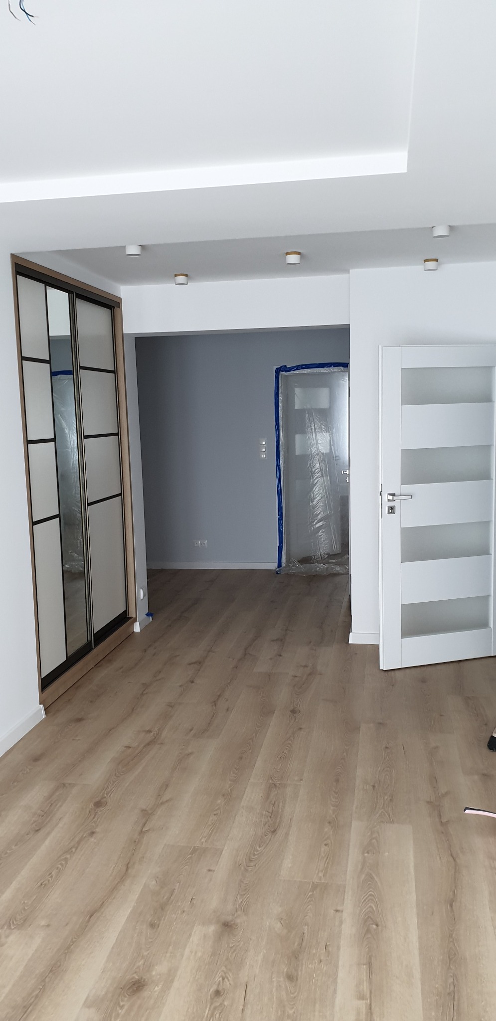 Jasne panele podłogowe ułożone w przestronnym wnętrzu z białą stolarką drzwiową i przesuwną szafą z czarnymi szprosami, ściany w odcieniach bieli i szarości, zabezpieczone drzwi folią ochronną.