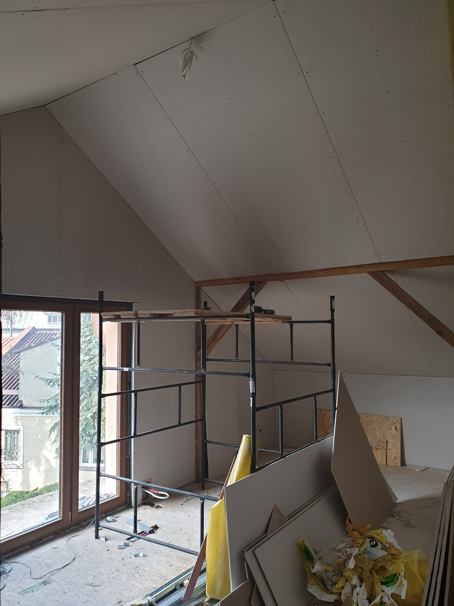 Poddasze w trakcie wykańczania płytami gipsowo-kartonowymi, widoczne rusztowanie, okno dachowe i materiały budowlane w nieładzie.