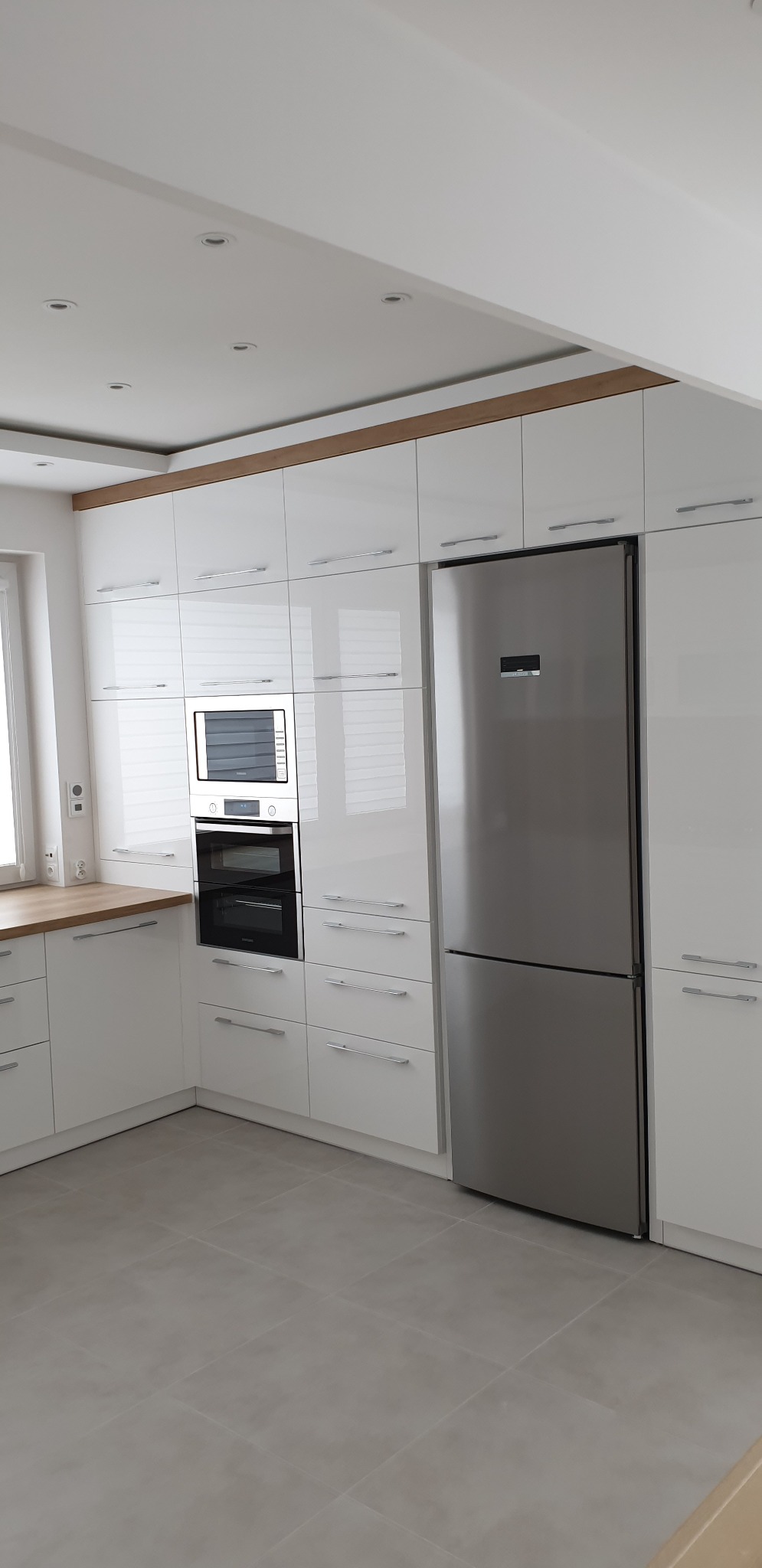 Nowoczesna, biała kuchnia z połyskiem w Ciechanowie, z zabudową pod sufit, srebrną lodówką i drewnianym akcentem na suficie.