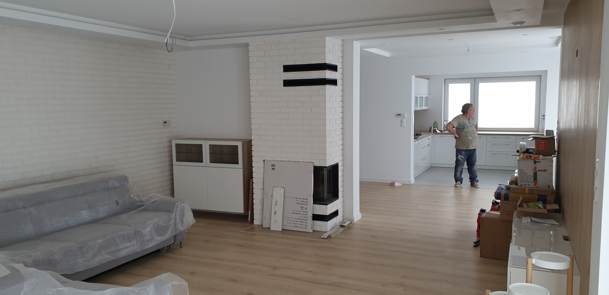 Wnętrze mieszkania w trakcie prac wykończeniowych: ściana z cegły pomalowanej na biało, obudowa kominka z białej cegły z czarnymi elementami dekoracyjnymi, meble częściowo zakryte folią ochronną...