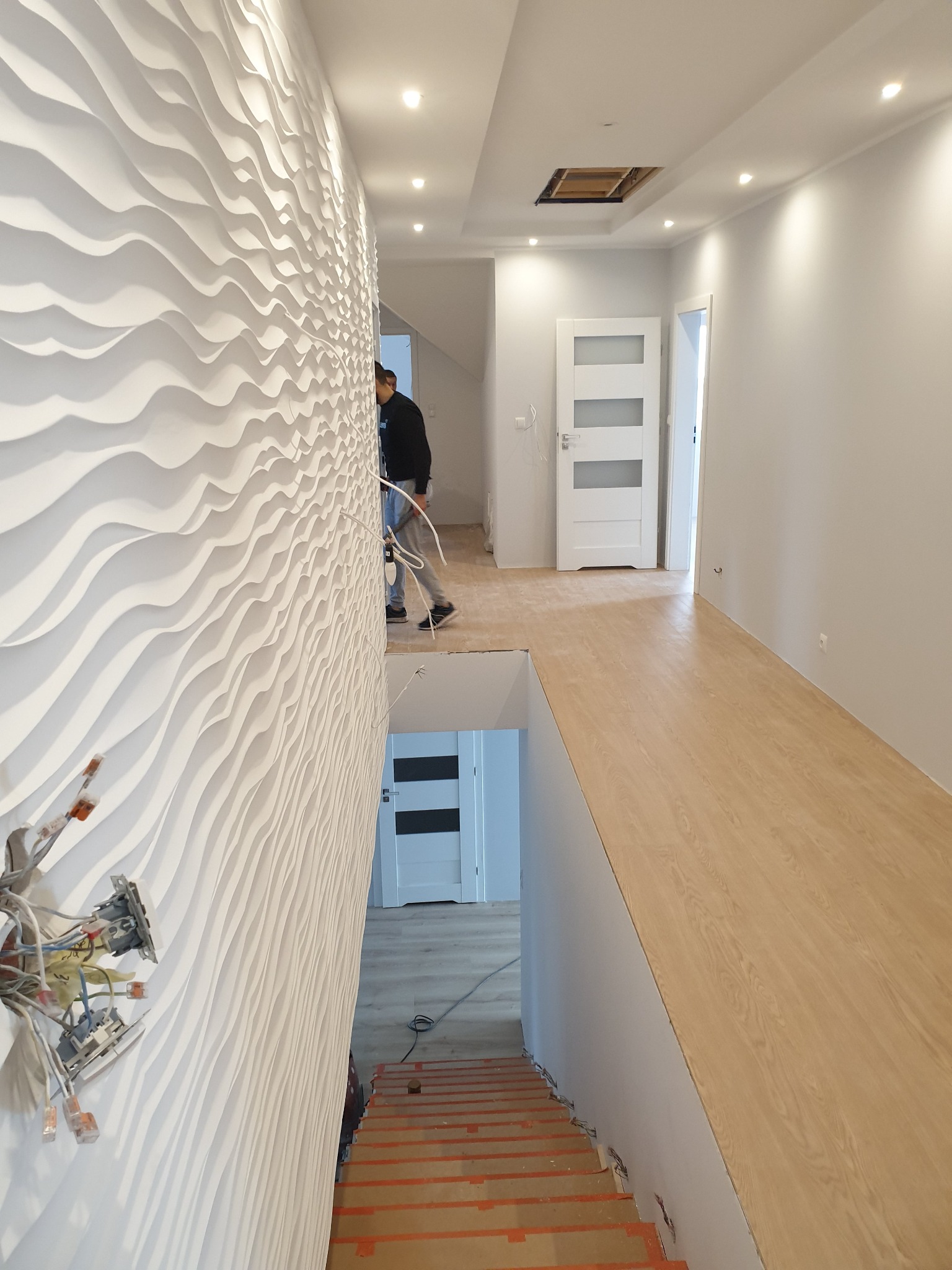 Korytarz w trakcie prac wykończeniowych: dekoracyjna ściana 3D z widoczną instalacją elektryczną, schody zabezpieczone folią, panele podłogowe i pracownik w tle.