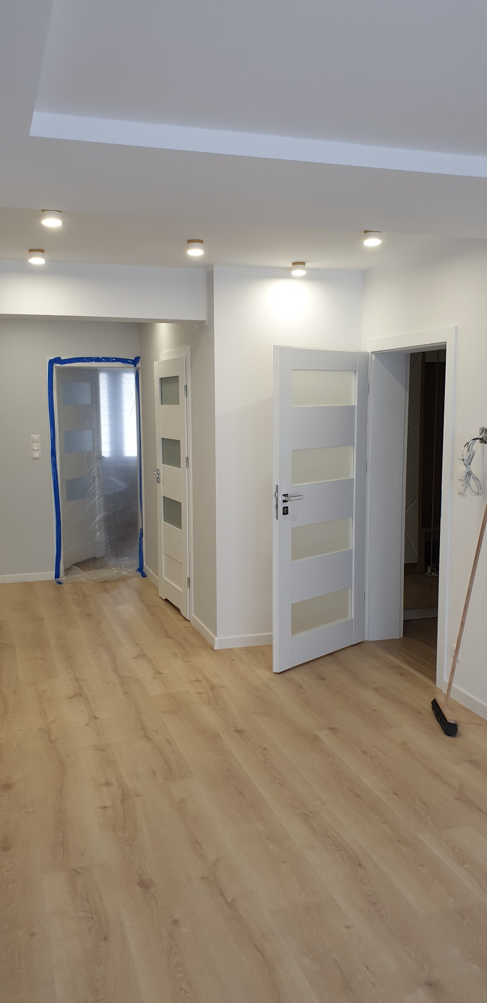 Wnętrze po odświeżeniu, z jasną podłogą, białymi ścianami i drzwiami z przeszkleniami; jedno z przejść zabezpieczone folią malarską, widoczna szczotka do zamiatania.
