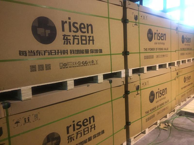 Palety z panelami słonecznymi marki RISEN, opakowane w kartonowe pudła z logo i opisami w języku chińskim, zabezpieczone taśmami i ustawione w magazynie.