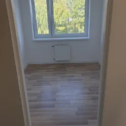 Wykończony pokój z panelami podłogowymi w odcieniach szarości i brązu, białe ściany, okno z widokiem na drzewa, biały grzejnik na ścianie, widok z korytarza przez białe futryny.