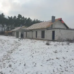 Dwa surowe budynki w stanie surowym otwartym, z częściowo położonym dachem z dachówki, otoczone śniegiem i lasem w tle, widoczny fragment osoby.