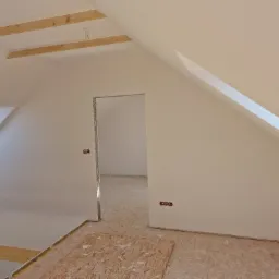 Poddasze po ociepleniu, z widoczną konstrukcją drewnianą, wygładzonymi ścianami, gniazdkami elektrycznymi i podłogą z płyt OSB przed dalszym wykończeniem.