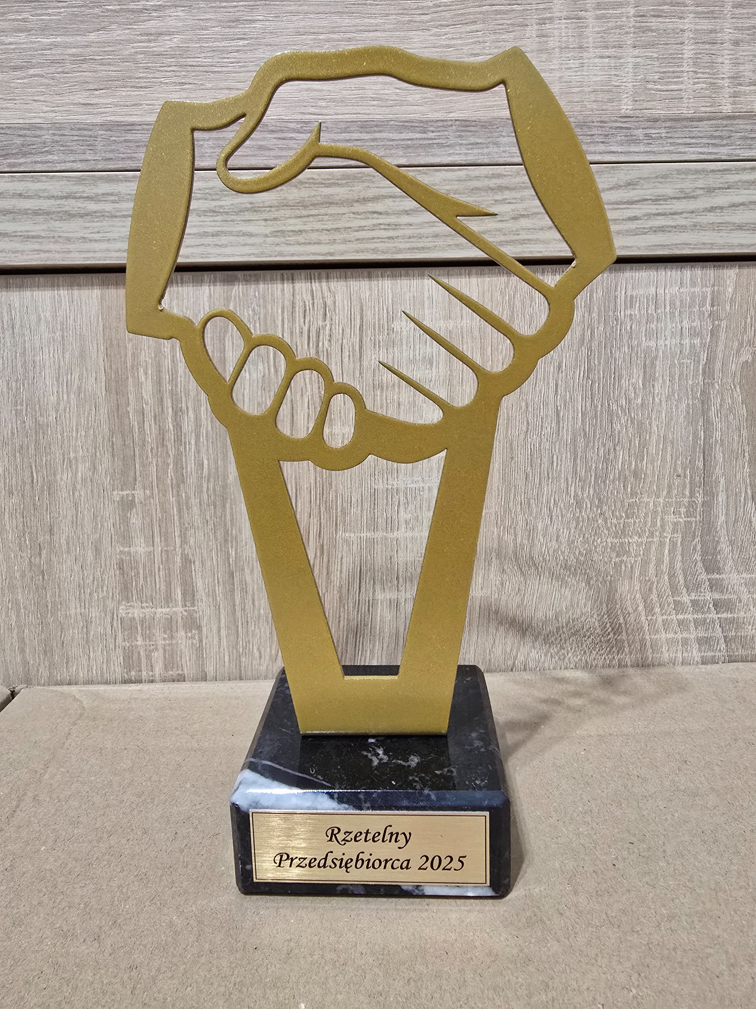 Złota statuetka z motywem uścisku dłoni na marmurowej podstawie z napisem 'Rzetelny Przedsiębiorca 2025' na tle drewnianej ściany.