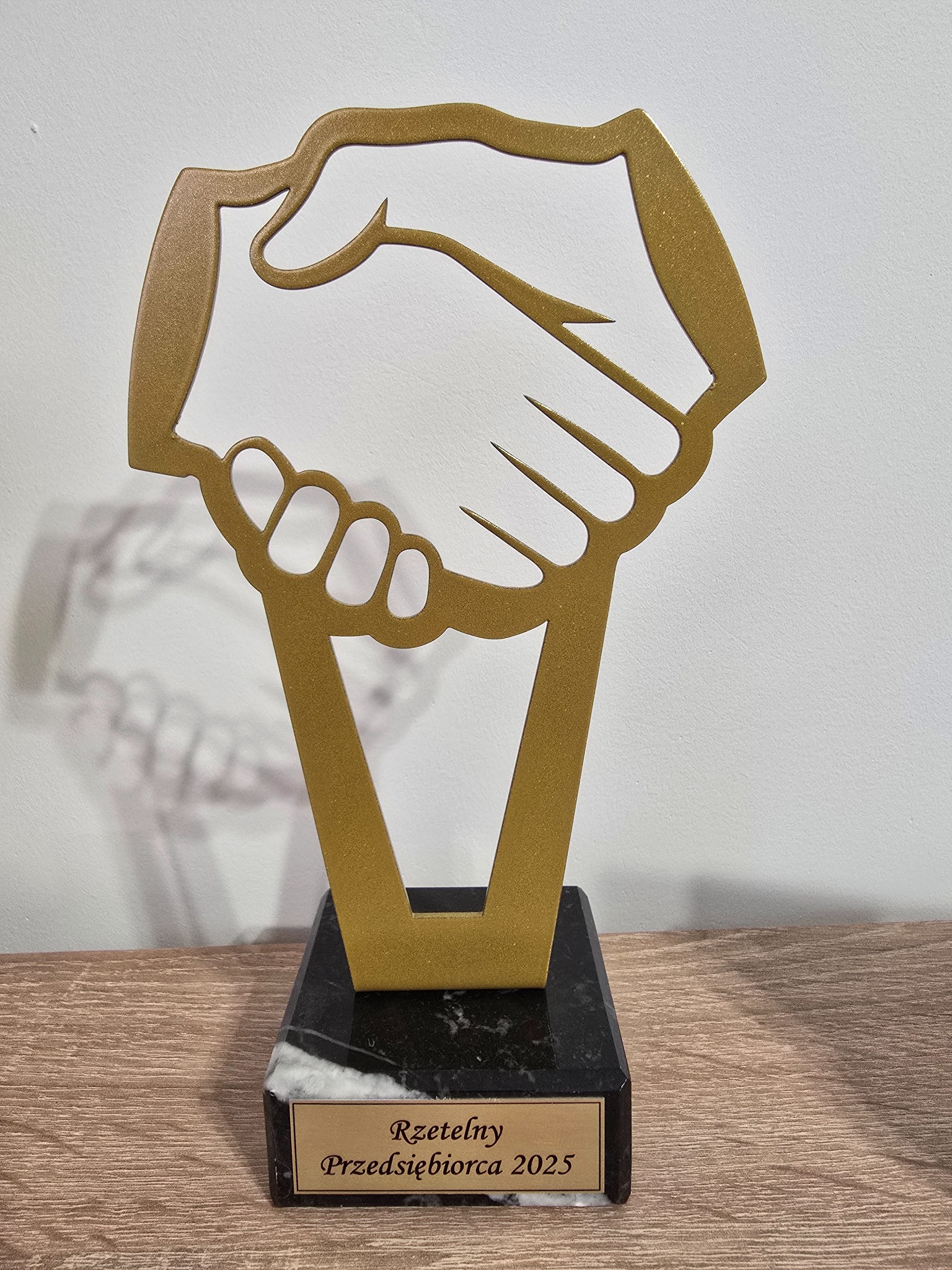 Złota statuetka z motywem uścisku dłoni na marmurowej podstawie z napisem 'Rzetelny Przedsiębiorca 2025' na drewnianym blacie, symbolizująca nagrodę.