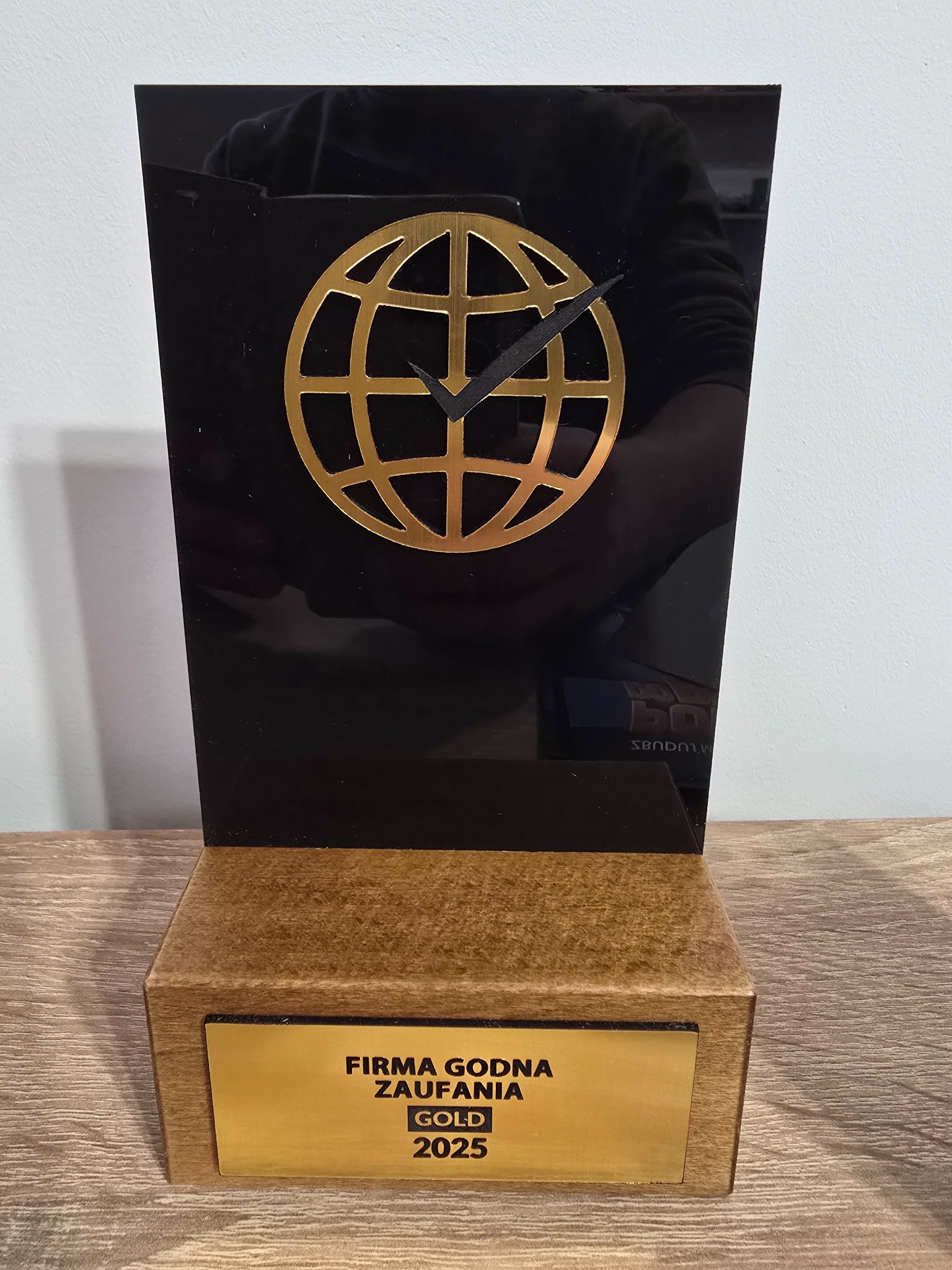 Statuetka 'Firma Godna Zaufania GOLD 2025' z czarnym akrylem i złotym globem-zegarem, na drewnianej podstawie, prezentowana na drewnianym blacie.
