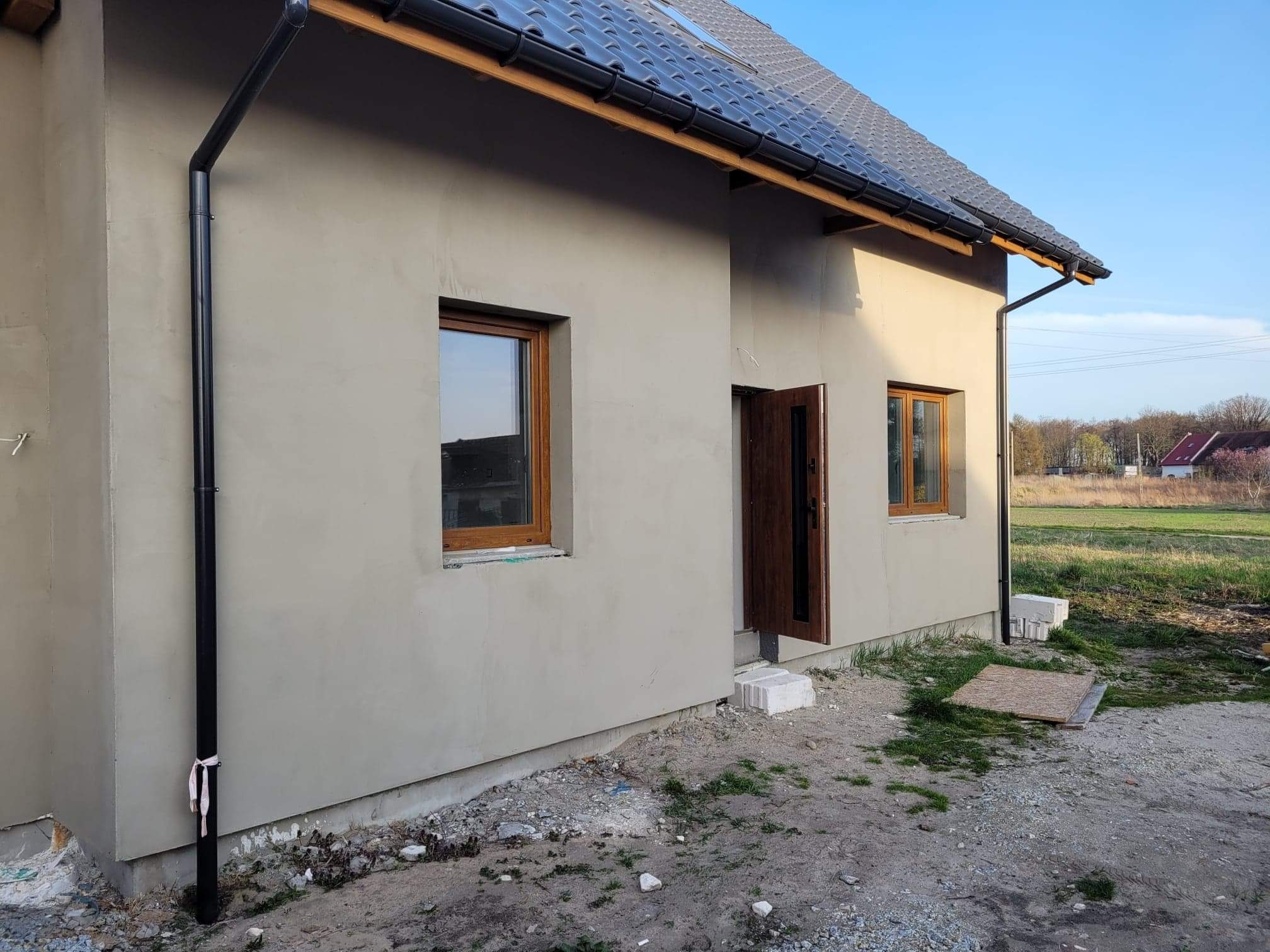 Surowa elewacja domu jednorodzinnego z brązowymi oknami i drzwiami, czarną rynną z zawiązaną szmatką, widok z zewnątrz na niezagospodarowany teren.