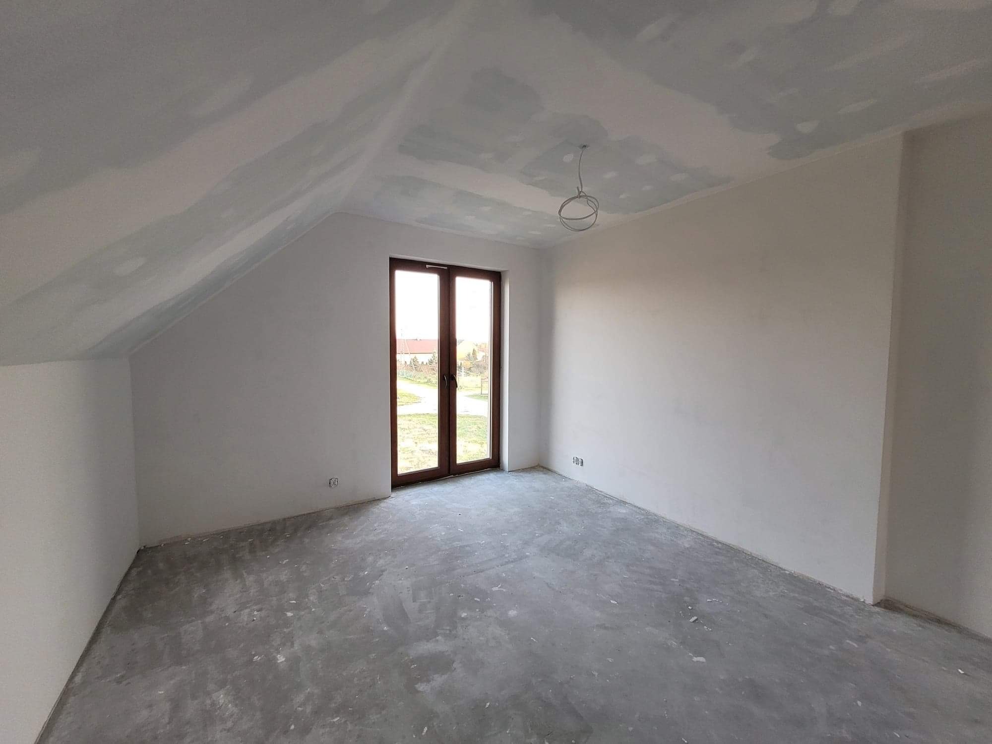 Poddasze w trakcie prac wykończeniowych: ściany i sufit pokryte warstwą szpachli, widoczne niedoskonałości i miejsca wymagające dalszej obróbki, surowy beton na podłodze, brak oświetlenia, okno...