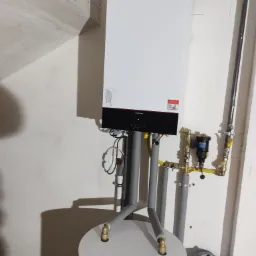 Nowoczesny piec grzewczy Viessmann z zasobnikiem, rurami w szarej izolacji i osprzętem, zamontowany przy białej ścianie w pomieszczeniu z widocznymi elementami konstrukcyjnymi.