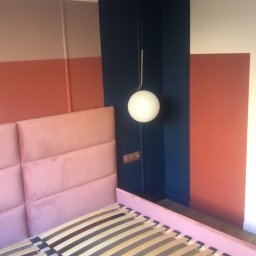 Daniel Mal Proff - Sypialnia w bloku: różowy zagłówek łóżka, drewniany stelaż, ściany w pasy (brąz, pomarańcz), granatowa wnęka z kulistą lampą. Nowoczesny design z akcentem kolorystycznym.