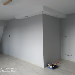 Daniel Mal Proff - Pokój po malowaniu w odcieniach szarości, z widocznymi elementami oświetlenia sufitowego i listwami przypodłogowymi. W rogu podłogi leżą narzędzia malarskie.