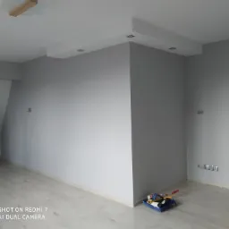 Pokój po malowaniu w odcieniach szarości, z widocznymi elementami oświetlenia sufitowego i listwami przypodłogowymi. W rogu podłogi leżą narzędzia malarskie.