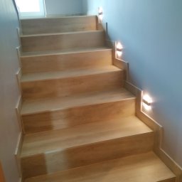 Daniel Mal Proff - Jasne, drewniane schody z oświetleniem punktowym na ścianie. Widok z dołu. Minimalistyczny design. W tle widoczne okno.