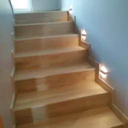 Jasne, drewniane schody z oświetleniem punktowym na ścianie. Widok z dołu. Minimalistyczny design. W tle widoczne okno.