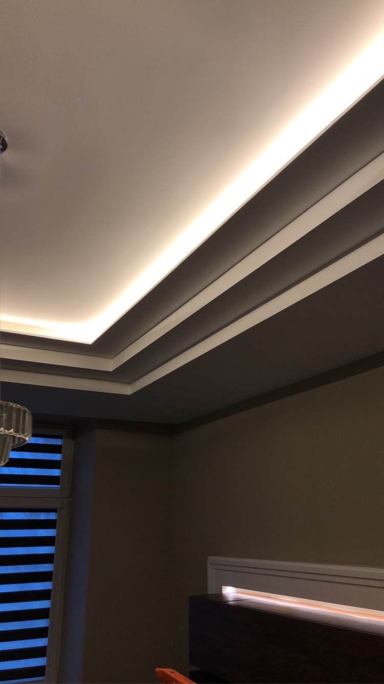 Nowoczesny sufit podwieszany z oświetleniem LED, listwy dekoracyjne i stonowana kolorystyka ścian. Widok z dołu, częściowo widoczna żaluzja okienna.