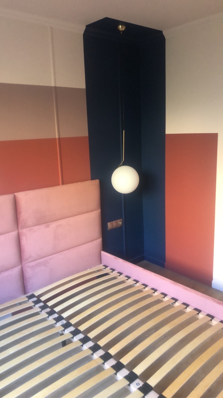 Sypialnia w bloku: różowy zagłówek łóżka, drewniany stelaż, ściany w pasy (brąz, pomarańcz), granatowa wnęka z kulistą lampą. Nowoczesny design z akcentem kolorystycznym.