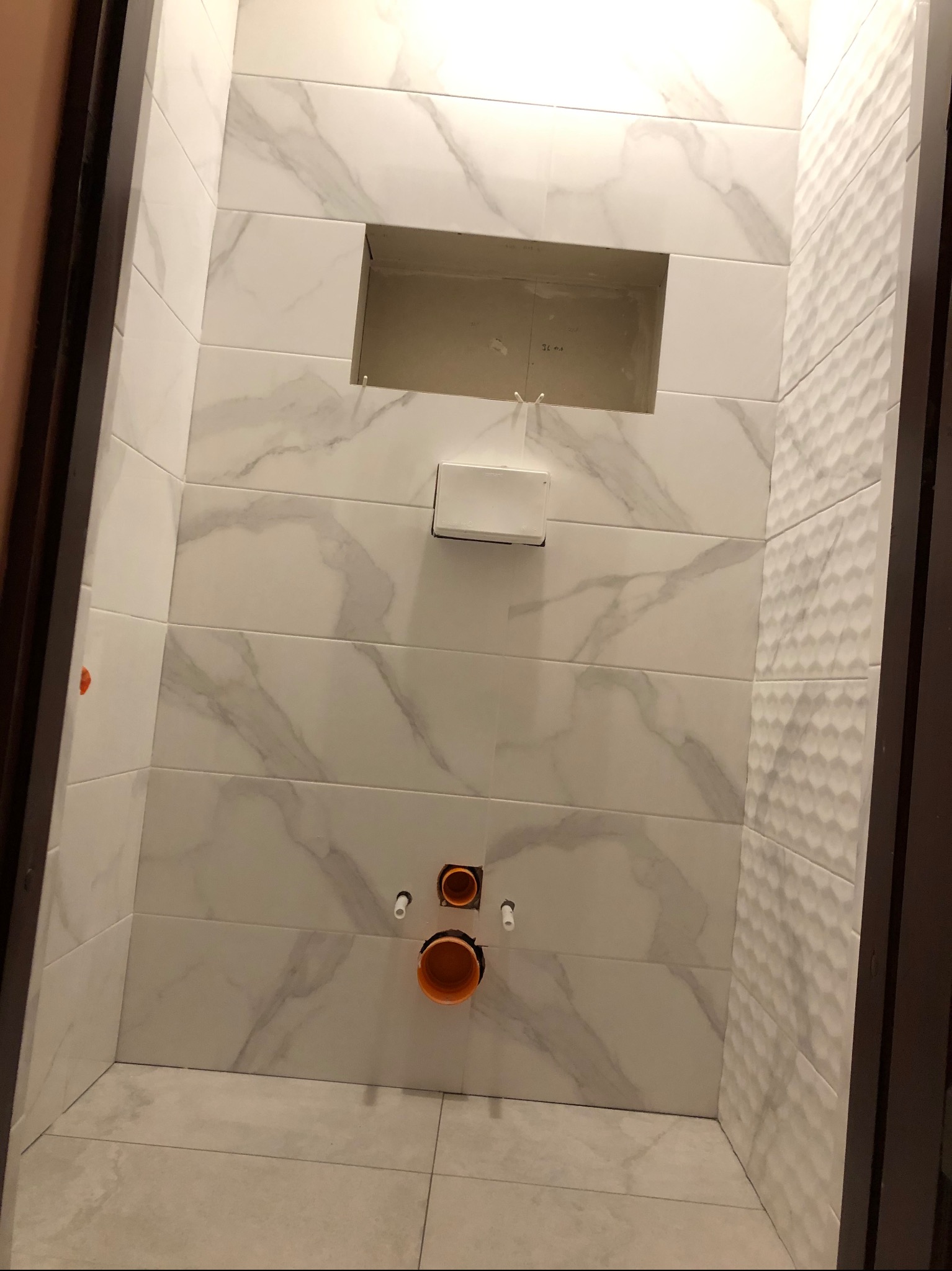 Wykończona łazienka z białymi płytkami imitującymi marmur, widoczna wnęka i instalacja wodna. Na ścianie płytki z fakturą 3D. Ujęcie z dołu.