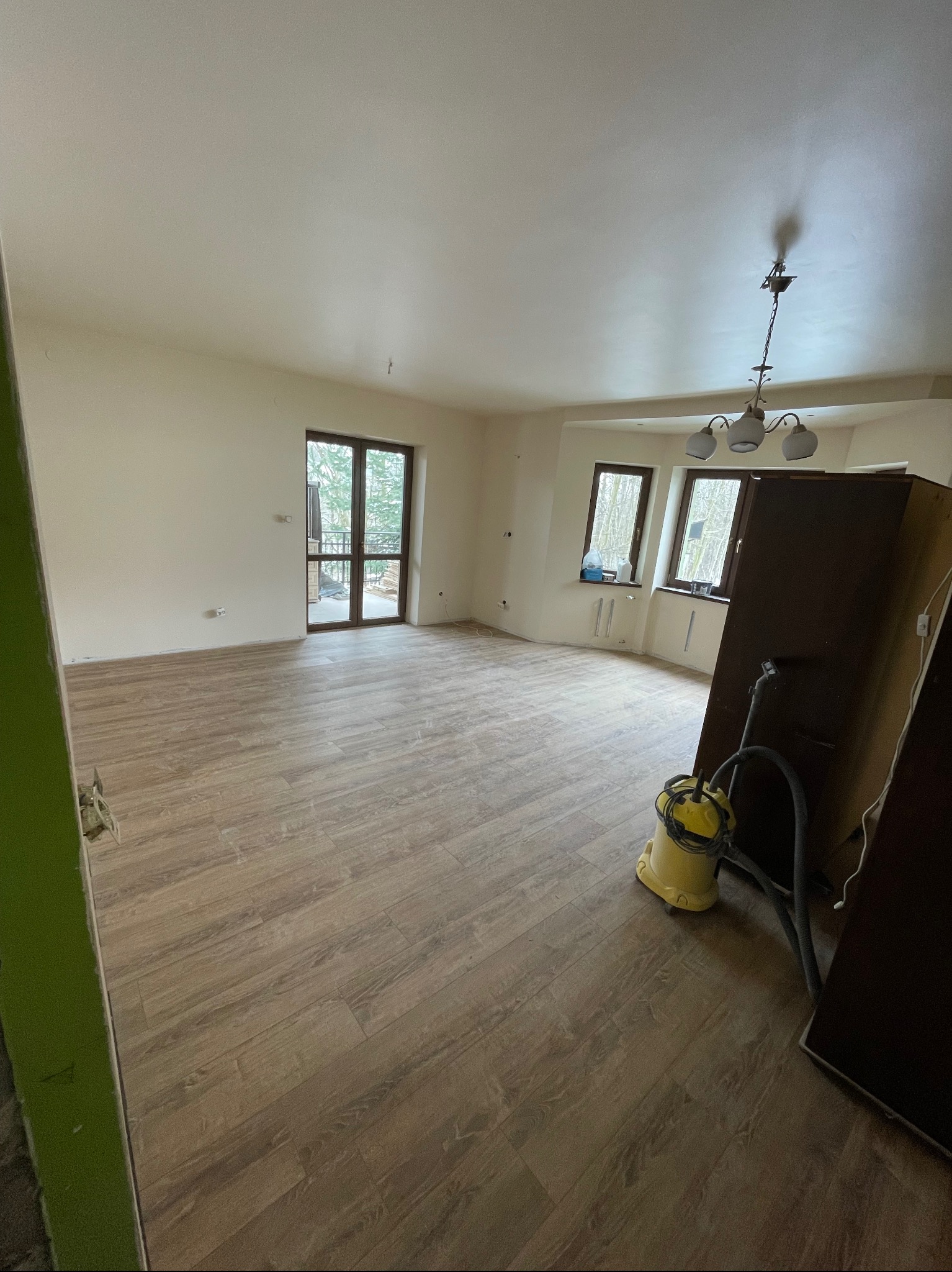 Jasne panele podłogowe w przestronnym pokoju z oknami i balkonem. Ściany przygotowane do malowania, widoczny odkurzacz i fragmenty mebli.