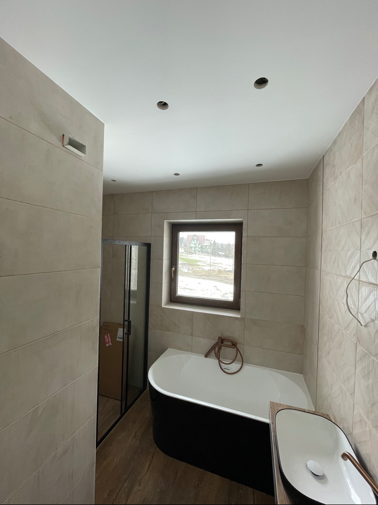 Jasna łazienka z czarną wanną wolnostojącą i kabiną prysznicową. Beżowe płytki na ścianach i drewnopodobna podłoga. Widoczne punkty oświetleniowe w suficie.