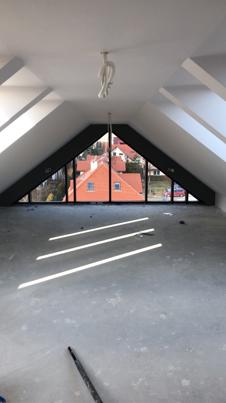 Poddasze z oknami dachowymi w kształcie trójkątów, surowe ściany i podłoga przygotowane do dalszych prac wykończeniowych. Widoczne promienie słońca i przewody oświetleniowe.