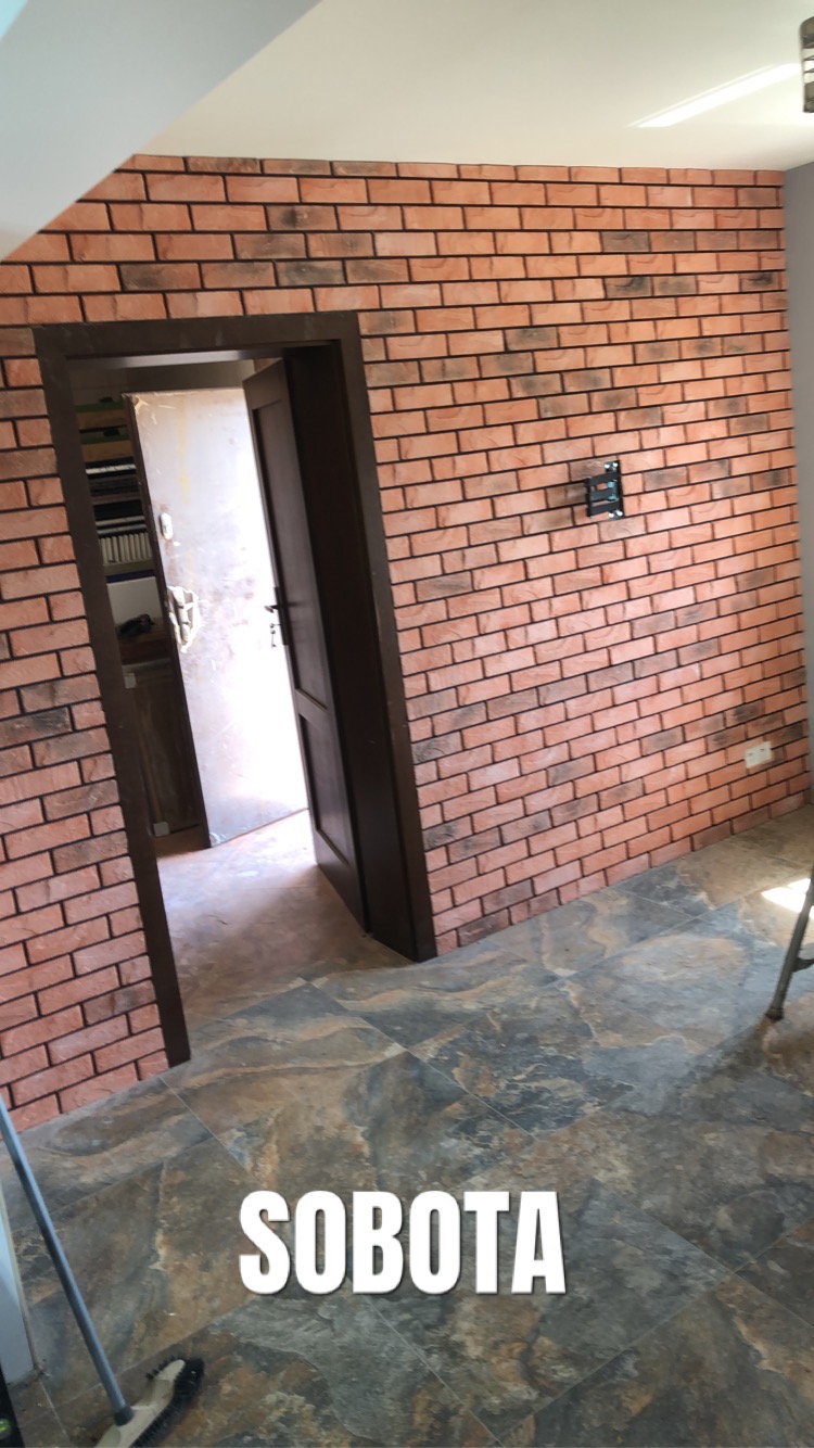 Ściana wykończona cegłą klinkierową w odcieniach czerwieni i brązu, z widocznym fragmentem podłogi z płytek imitujących kamień i otwartymi drzwiami.