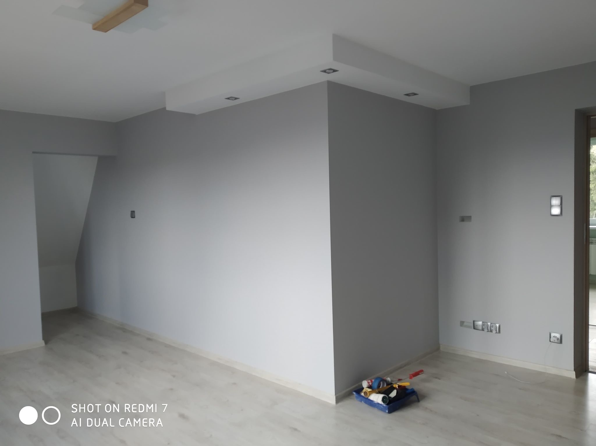 Pokój po malowaniu w odcieniach szarości, z widocznymi elementami oświetlenia sufitowego i listwami przypodłogowymi. W rogu podłogi leżą narzędzia malarskie.