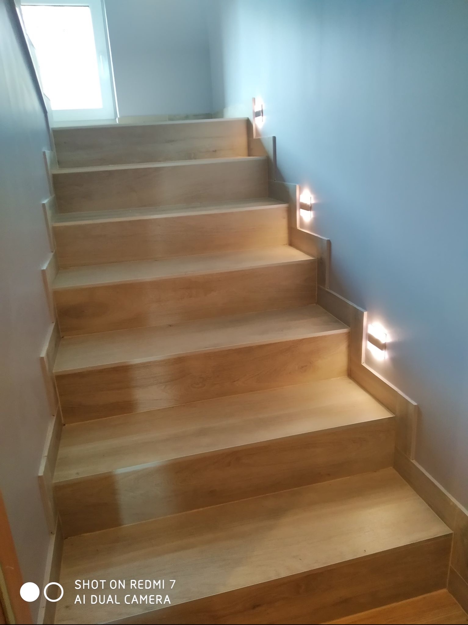 Jasne, drewniane schody z oświetleniem punktowym na ścianie. Widok z dołu. Minimalistyczny design. W tle widoczne okno.