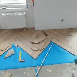 Układanie jasnych paneli podłogowych w jodełkę na niebieskim podkładzie, widoczne narzędzia: miarka, nóż i kątownik.