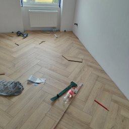 MaxBud - Układanie parkietu w jodełkę francuską, widoczne narzędzia, niedopita butelka Coca-Coli, ołówek stolarski i fragment instrukcji, w tle okno z widokiem na budynek.